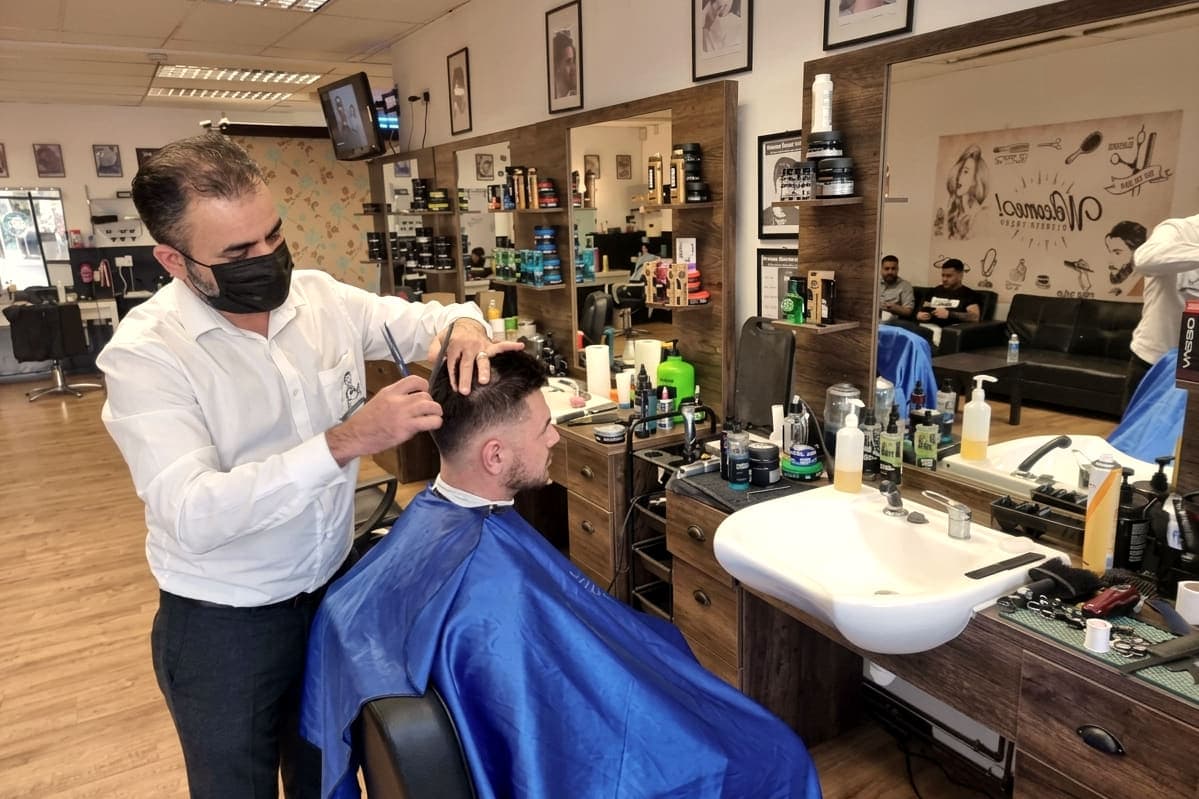 A barber in a mask cutting a client's hair with scissors over comb at the styling chair