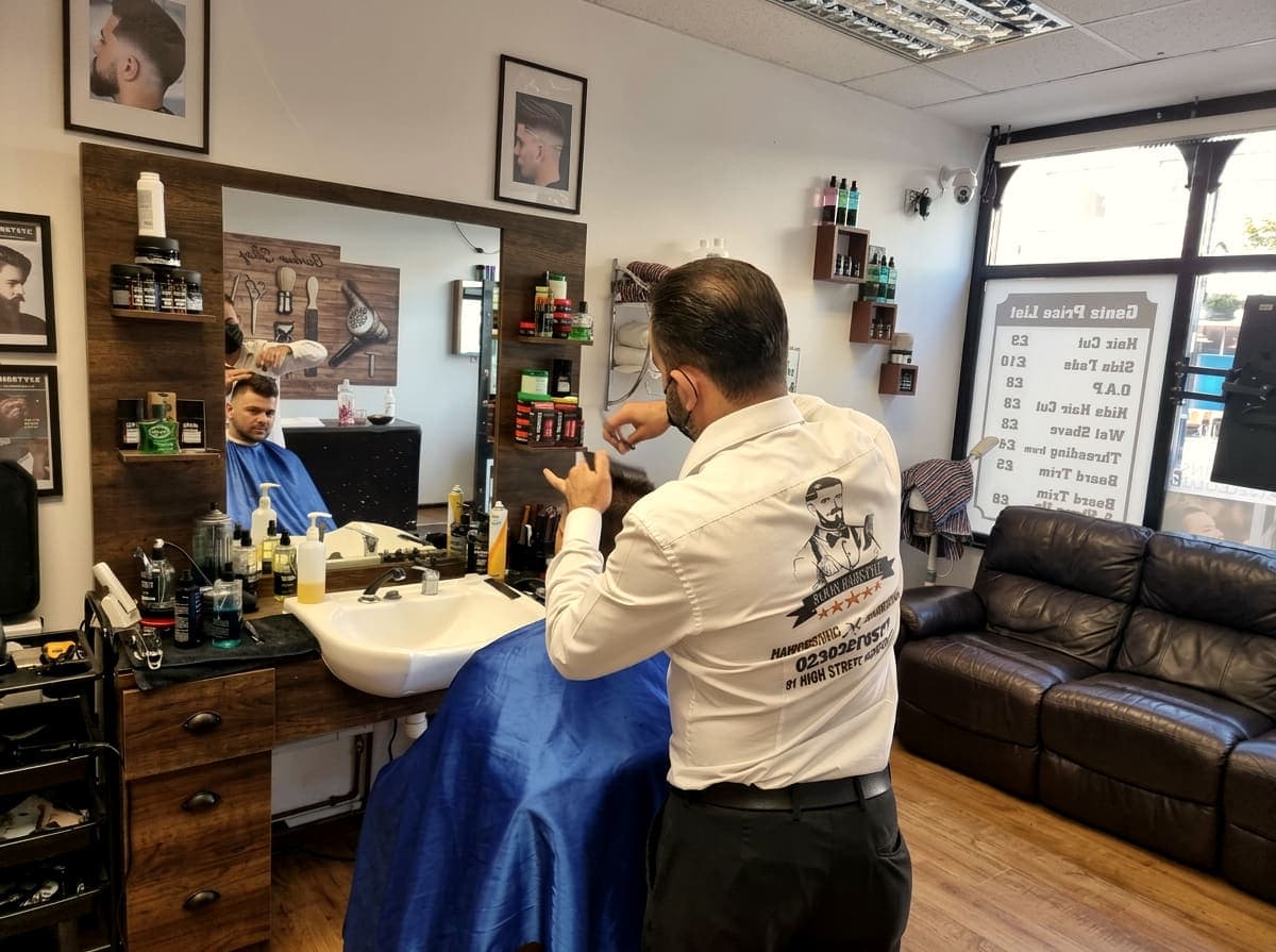 A barber working at a mirrored station beside the basin, with the gents price list on the window behind